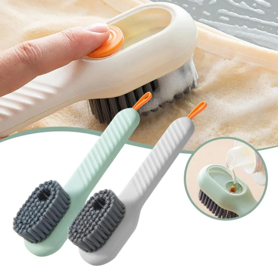 Cepillo de limpieza con dispensador de jabón en uso sobre tela, acompañado de dos unidades adicionales en colores verde menta y blanco, con cerdas oscuras y mango ergonómico.