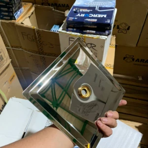 Ducha cuadrada en acero inoxidable sin tubo, con acabado brillante y orificio central dorado, mostrada en mano sobre fondo de cajas de almacenamiento.