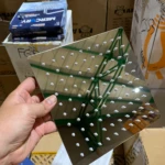 Ducha cuadrada en acero inoxidable sin tubo, con múltiples orificios para chorro uniforme, mostrada en mano sobre cajas de almacenamiento.