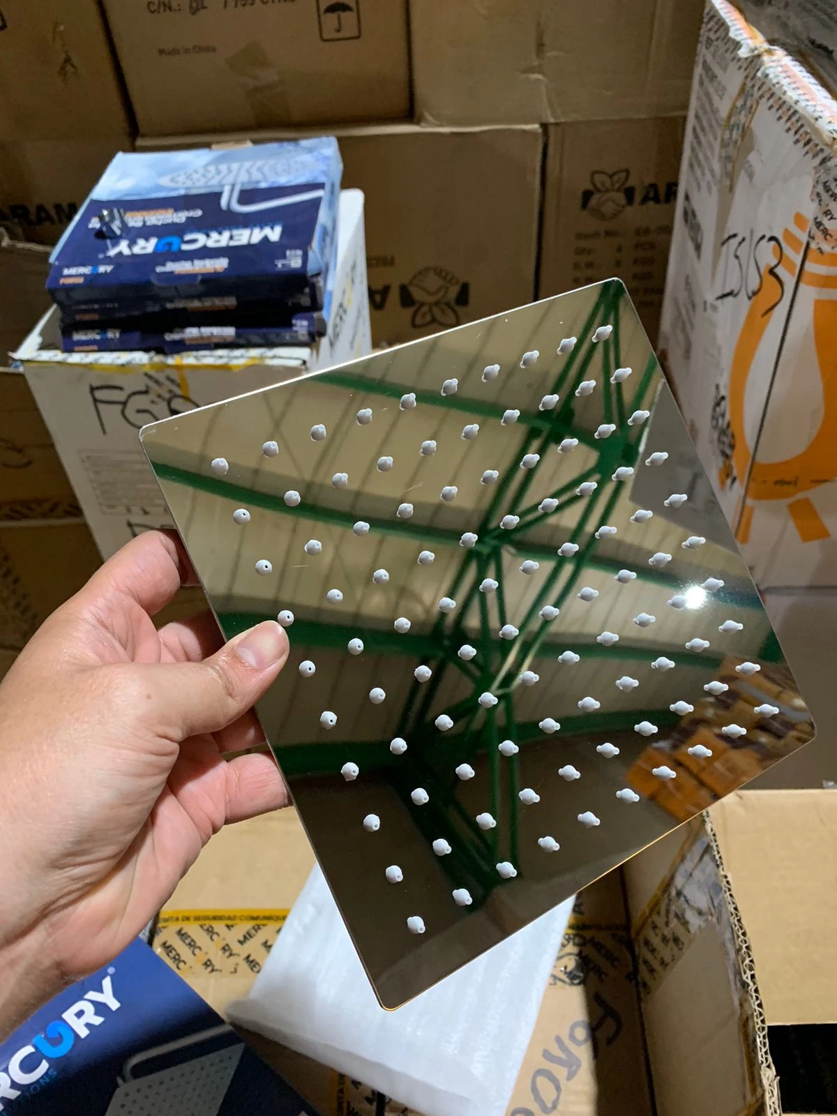 Ducha cuadrada en acero inoxidable sin tubo, con múltiples orificios para chorro uniforme, mostrada en mano sobre cajas de almacenamiento.