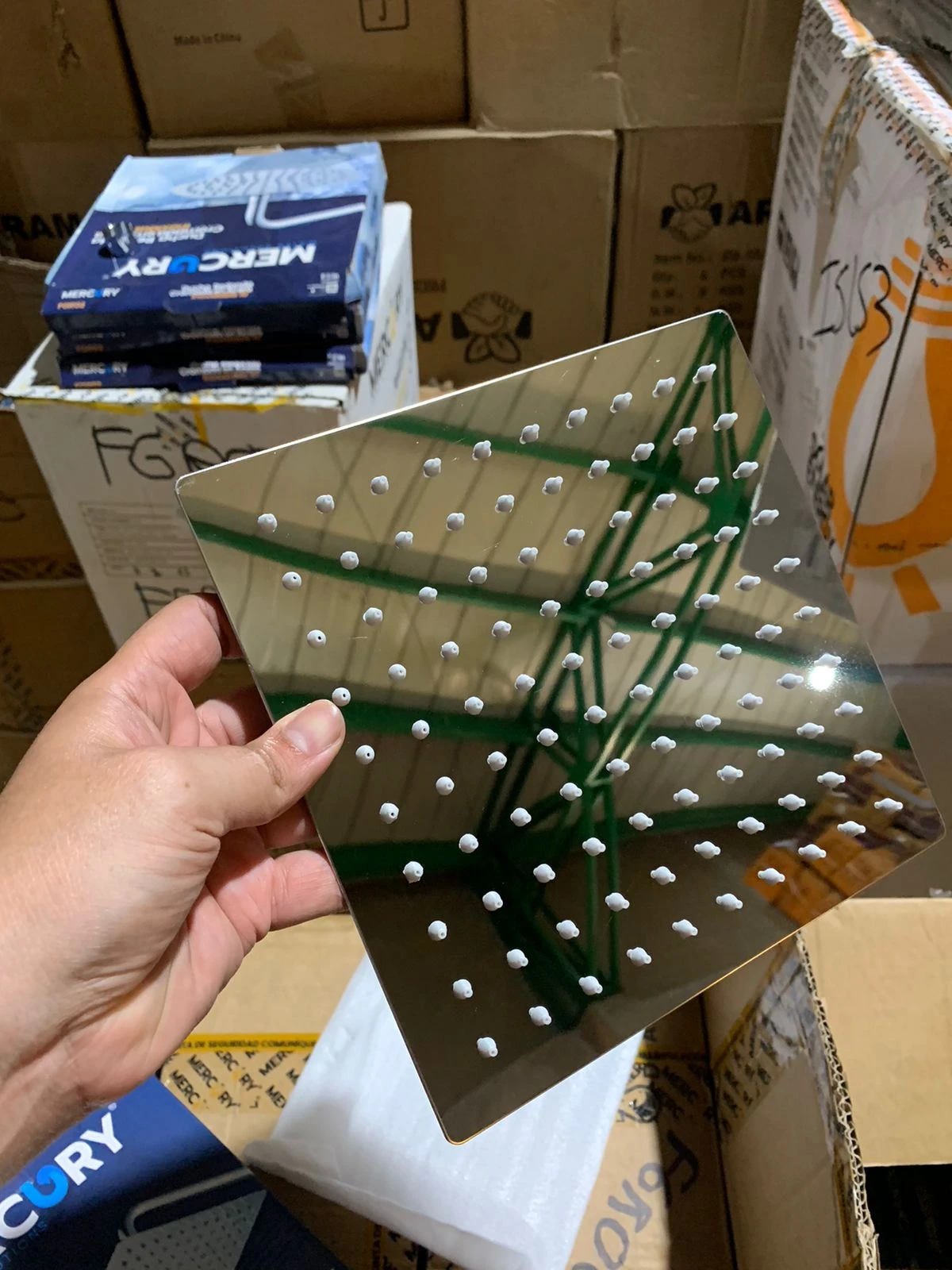 Ducha cuadrada en acero inoxidable sin tubo, con múltiples orificios para chorro uniforme, mostrada en mano sobre cajas de almacenamiento.