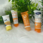 Cuatro tubos de crema solar SPF 65 de Kaliya Beauty, en tonos naranjas y blancos, dispuestos sobre superficie blanca con follaje verde al fondo.