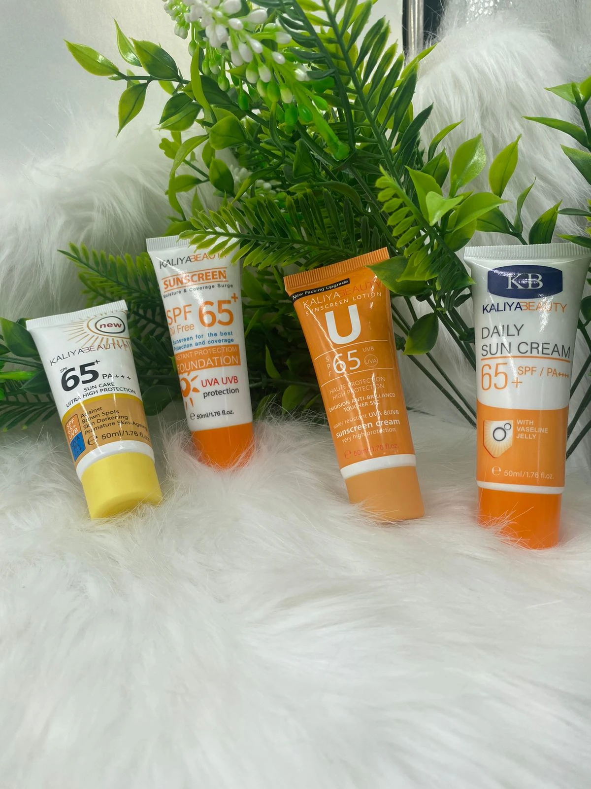 Cuatro tubos de crema solar SPF 65 de Kaliya Beauty, en tonos naranjas y blancos, dispuestos sobre superficie blanca con follaje verde al fondo.