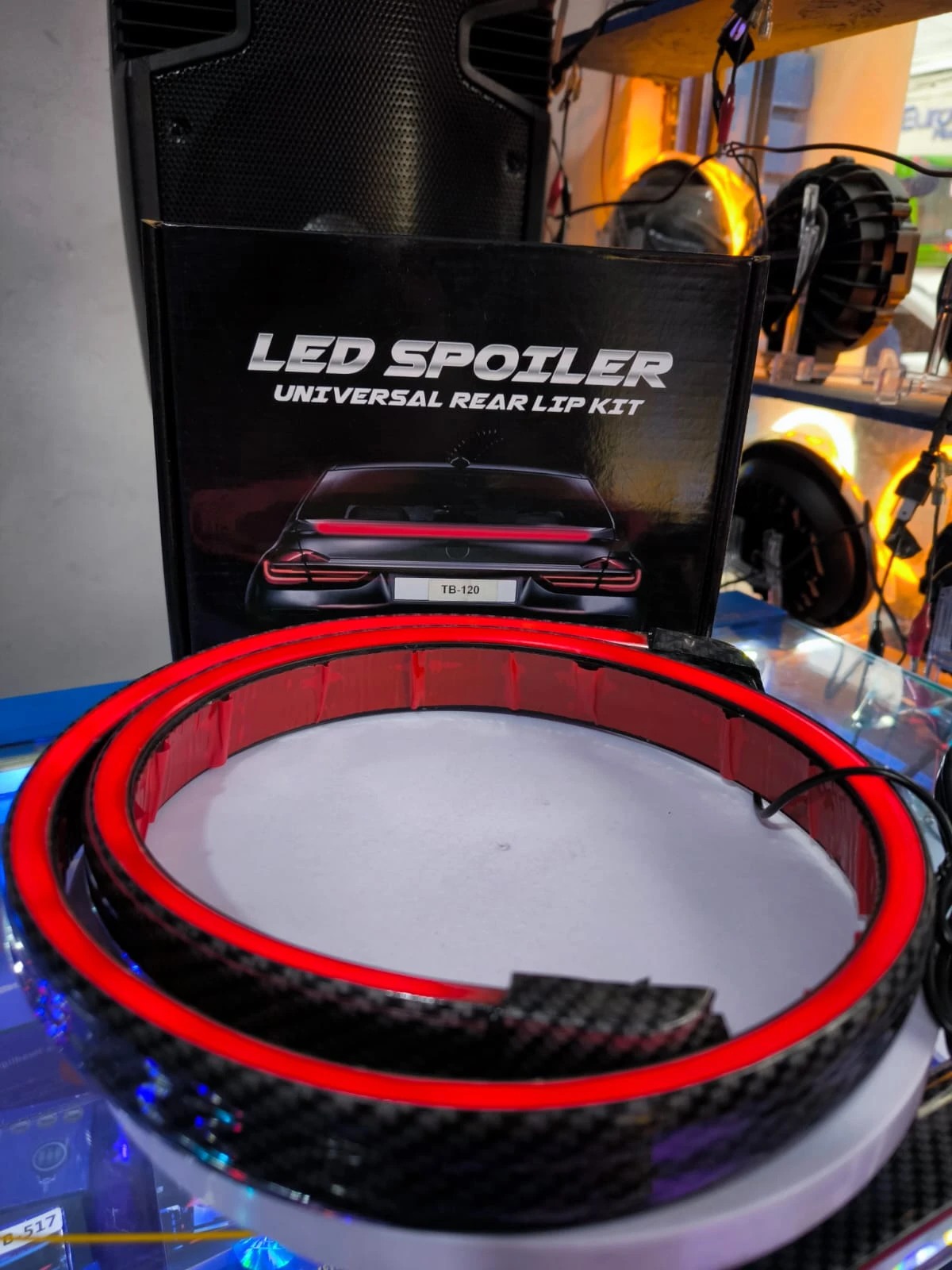 Spoiler trasero con luces LED rojas y acabado en fibra de carbono, presentado sobre base blanca junto a su caja de venta al por menor.