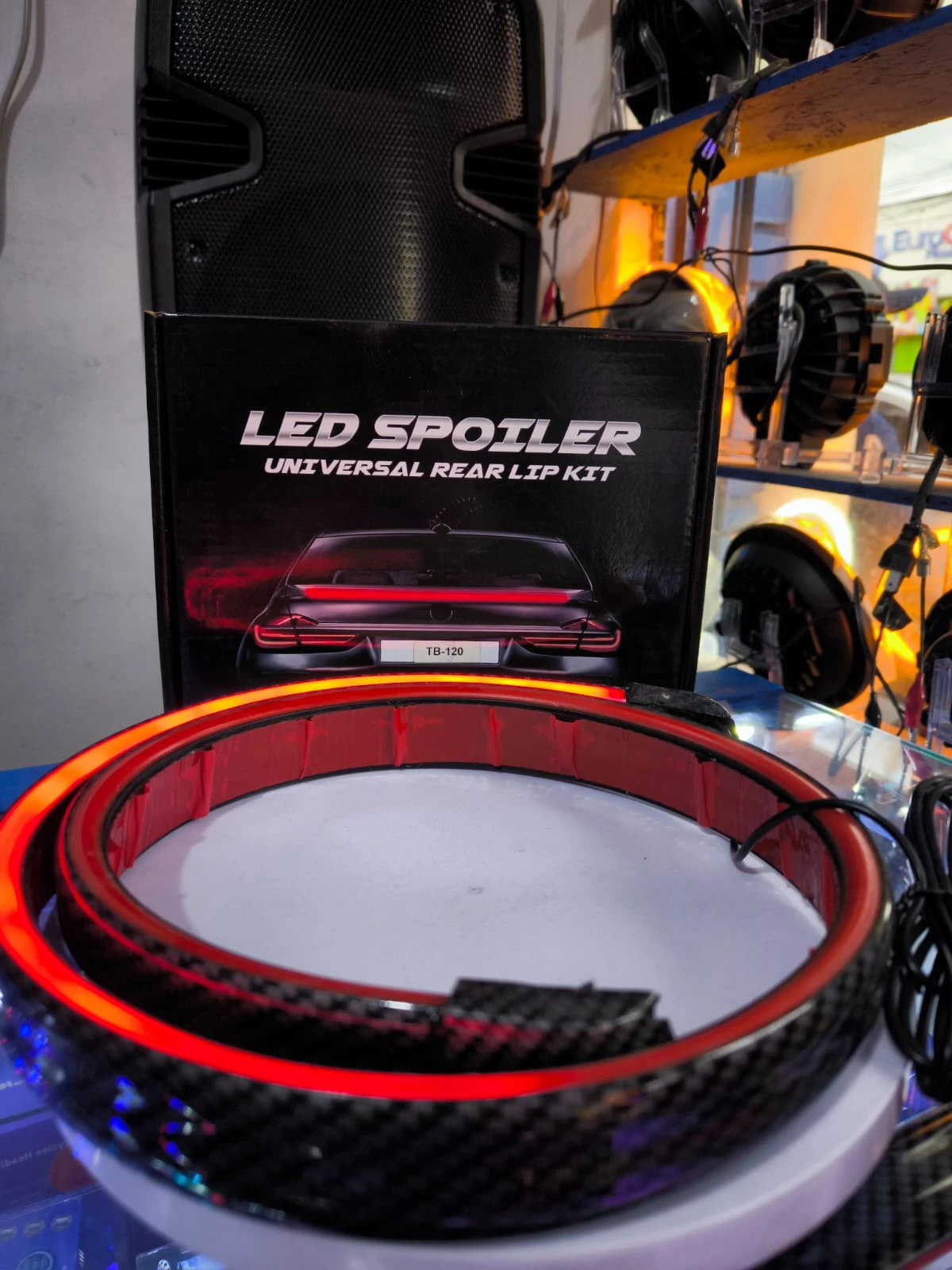 Spoiler trasero de fibra de carbono con luces LED rojas, montado sobre base blanca, junto a su caja original que indica "LED Spoiler Universal Rear Lip Kit".