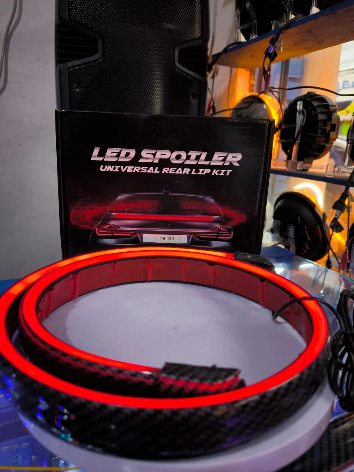Anillo de luces LED rojas con acabado fibra de carbono, montado sobre base blanca, junto a su caja negra que muestra el texto "LED SPOILER UNIVERSAL REAR LIP KIT" y una del producto instalado en un vehículo.