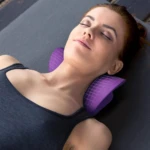Mujer recostada sobre almohada ergonómica de tracción cervical en color morado, con diseño de rejilla y forma envolvente para soporte de cuello y hombros.