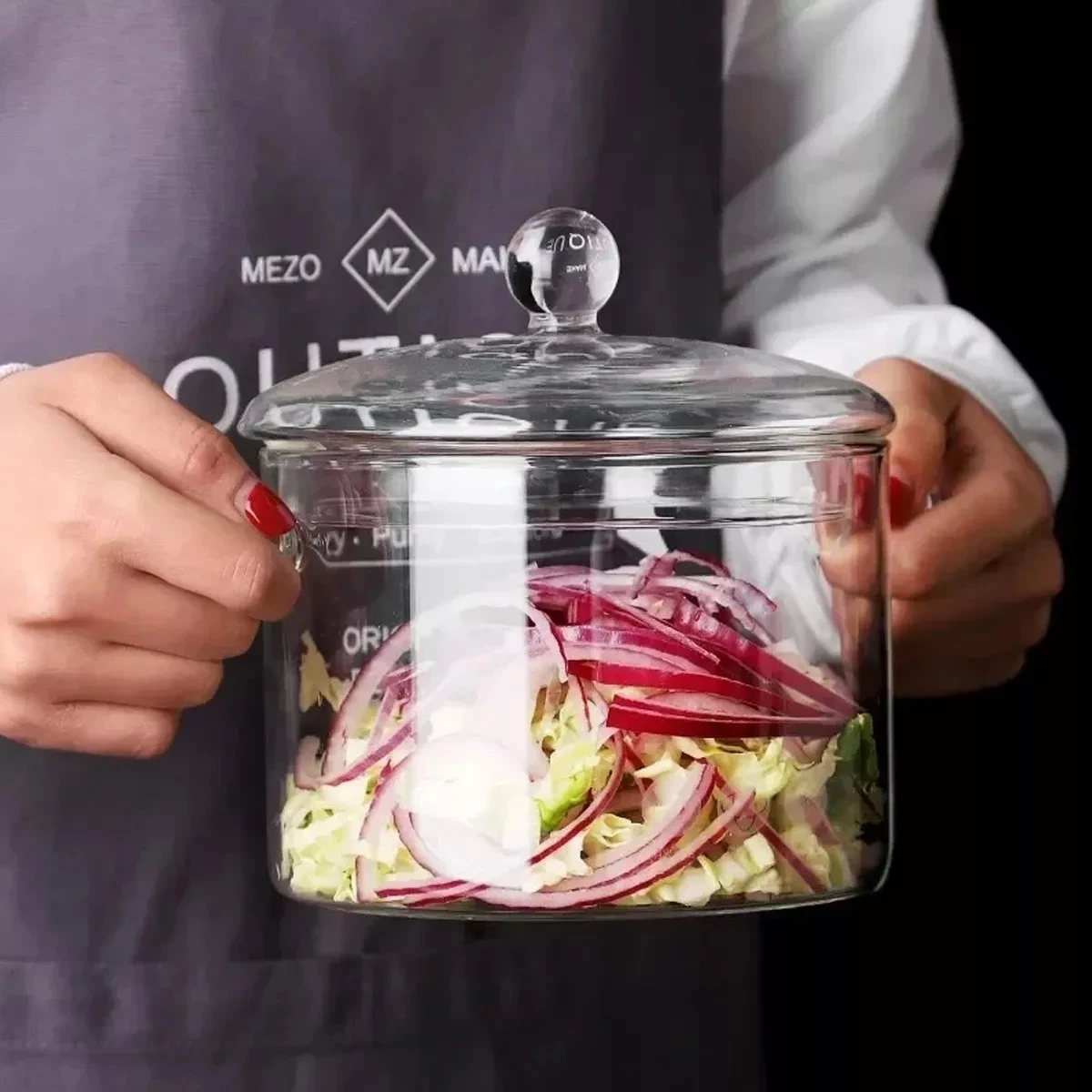 Olla de vidrio borosilicato transparente con tapa de cristal y mango esférico, conteniendo cebolla morada y lechuga, sostenida por manos con uñas pintadas de rojo.