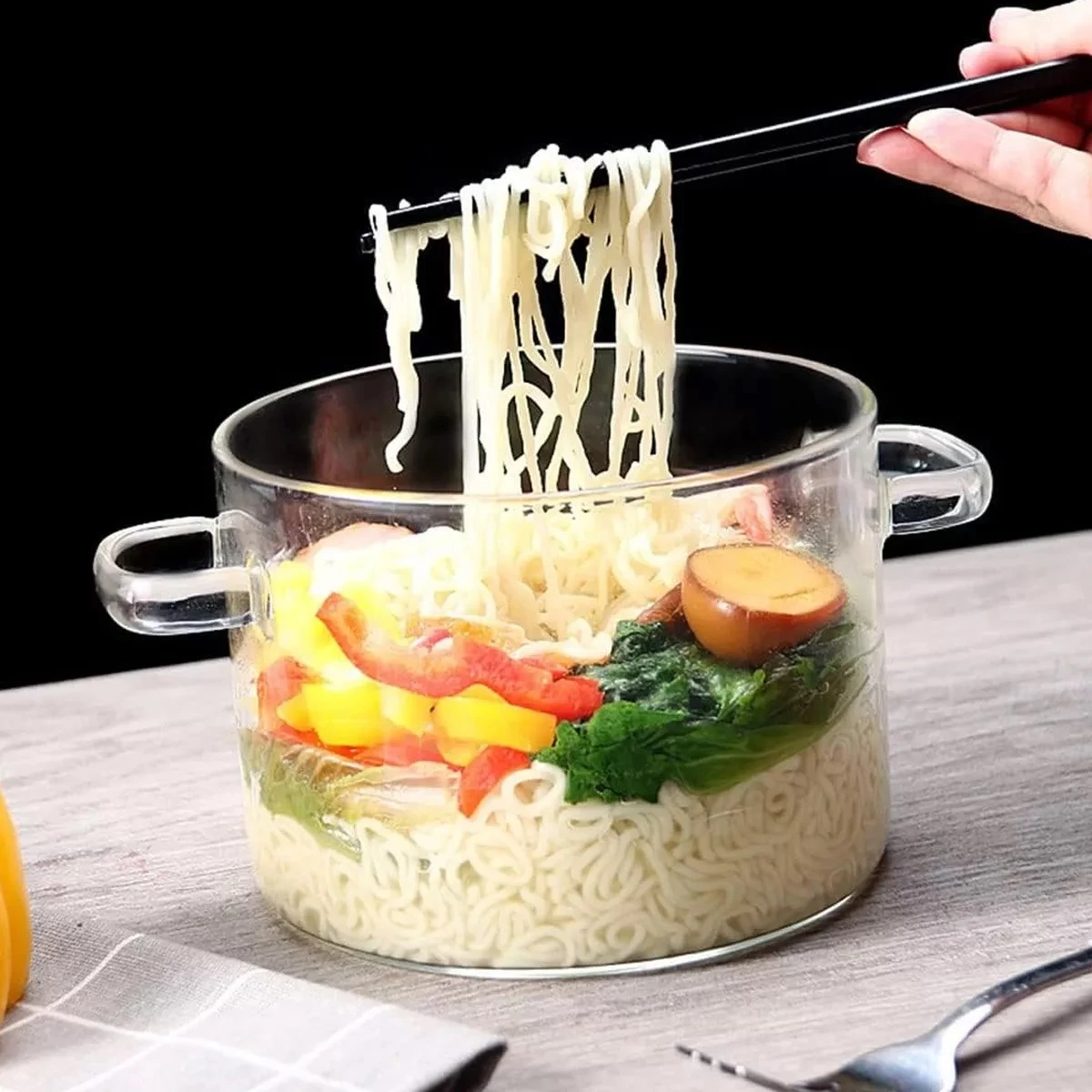 Olla de vidrio transparente con asas, mostrando pasta cocida, vegetales y huevo al vapor, siendo servida con palillos sobre mesa de madera.
