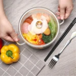Olla de vidrio borosilicato con tapa transparente, mostrando vegetales y huevo cocinándose; manos sujetan asas, acompañada de tenedor, palillos y pimiento amarillo sobre mesa.