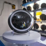 Farola H4 de aluminio para camiones, vista frontal con anillo LED blanco y lente oscura, montada sobre pedestal blanco en exhibición.