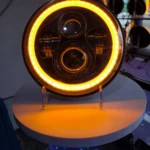 Farola H4 de aluminio para camiones, vista frontal con anillo LED encendido en tono ámbar, montada sobre soporte blanco sobre mesa.