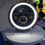 Farola H4 de aluminio para camiones, vista frontal con anillo LED encendido y lentes centrales visibles sobre soporte blanco.