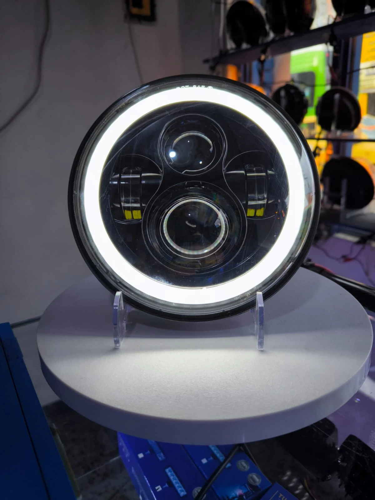 Farola H4 de aluminio para camiones, vista frontal con anillo LED encendido y lentes centrales visibles sobre soporte blanco.