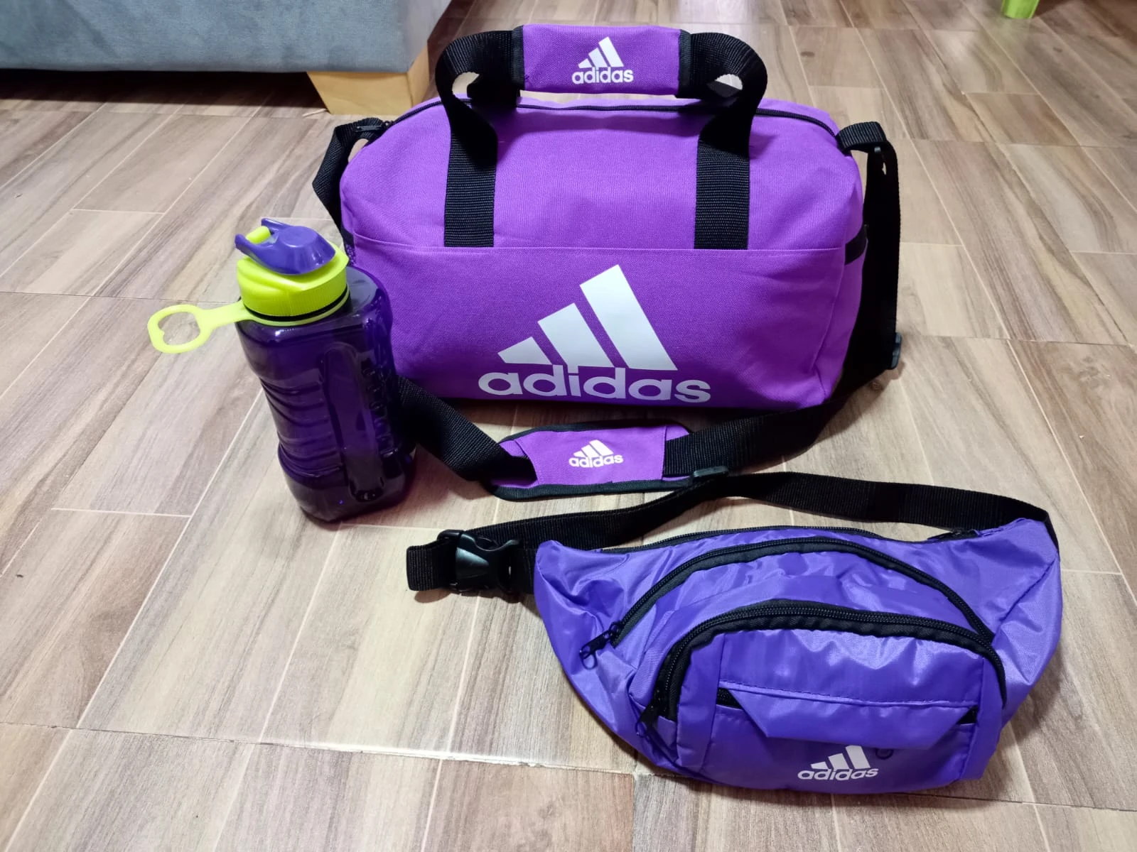 Bolsa deportiva morada Adidas con botiloto de 900 ml y faja de cintura, sobre piso de madera.