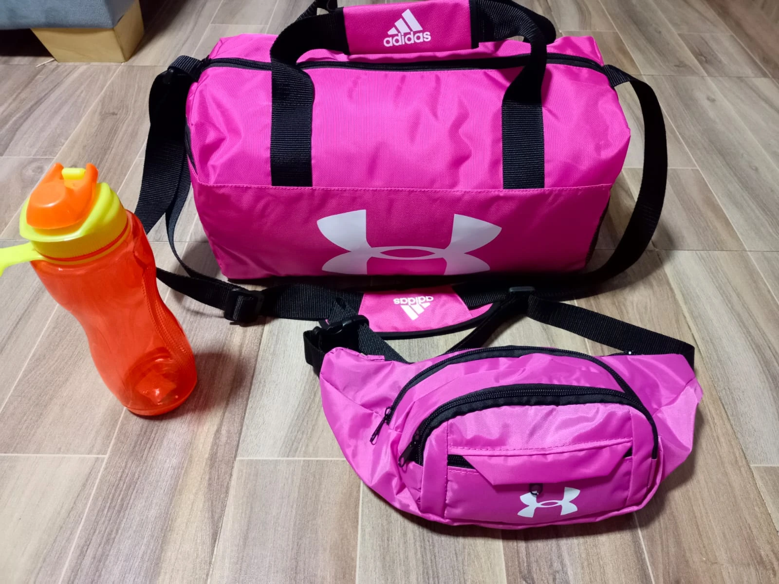 Bolsa deportiva rosa con logo de Under Armour, acompañada de una botella naranja de 900 ml y un fanny pack del mismo color, sobre piso de madera.