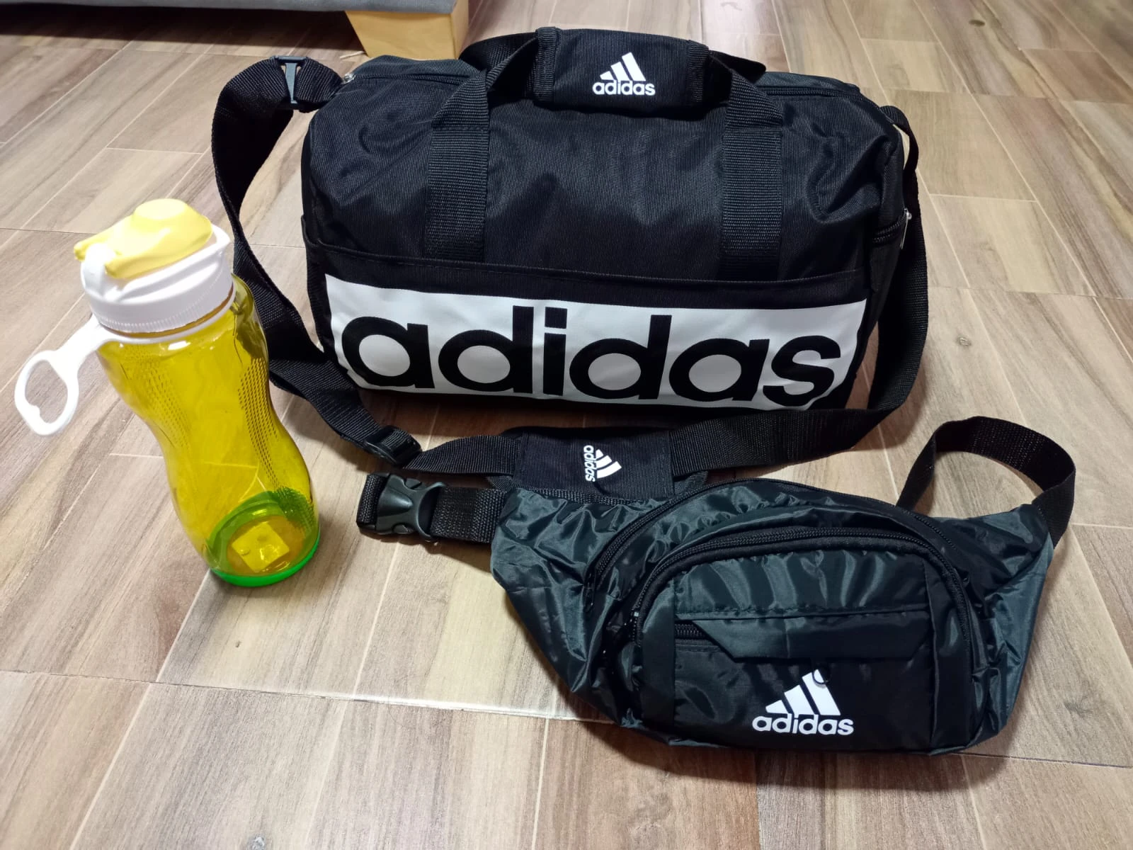 Bolsa deportiva negra con logo Adidas, acompañada de botiloto amarillo de 900 ml y faja negra con estampado camuflado, sobre piso de madera.
