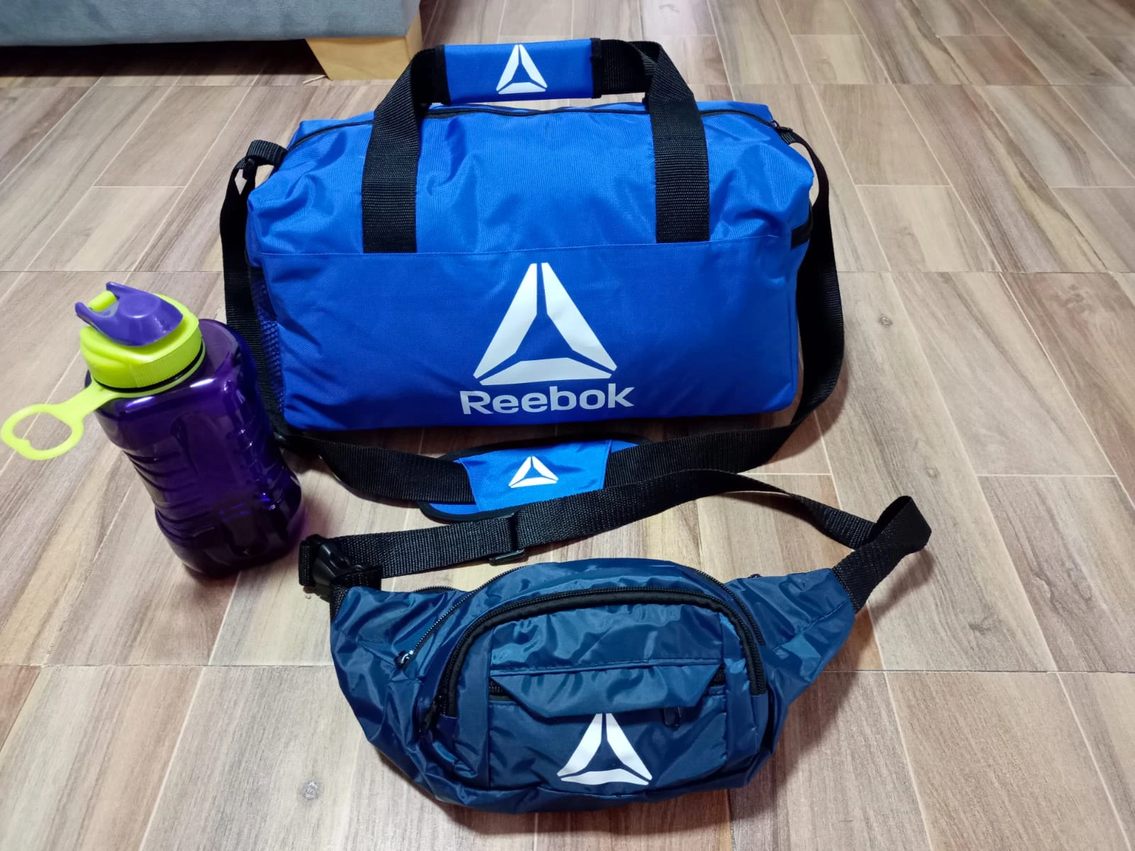Bolsa deportiva azul con logo Reebok, botiloto morado de 900 ml y faja azul oscura, sobre piso de madera.