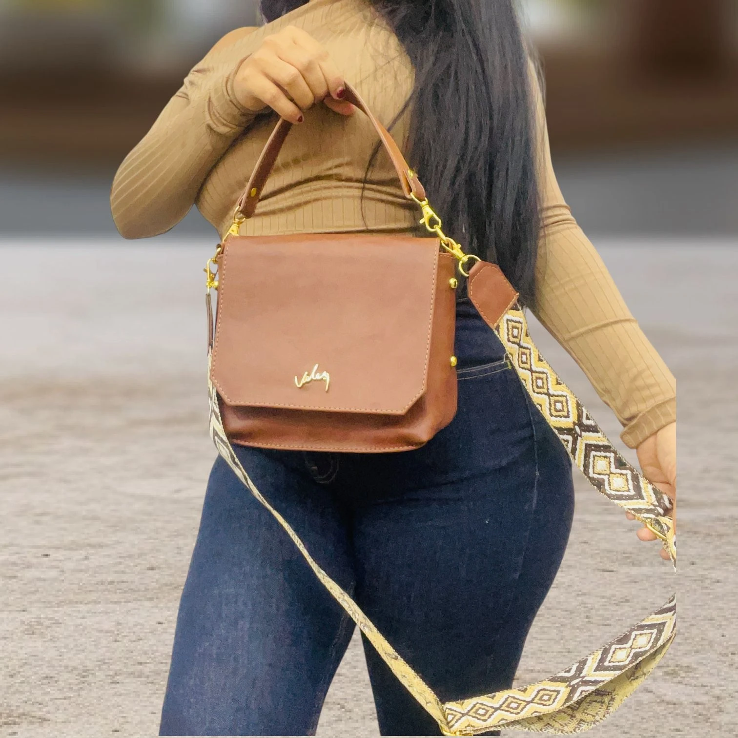 Carriel Dacota Dama Velez en cuero sintético marrón, con asa corta y correa larga estampada, usado por mujer con jeans y top beige.