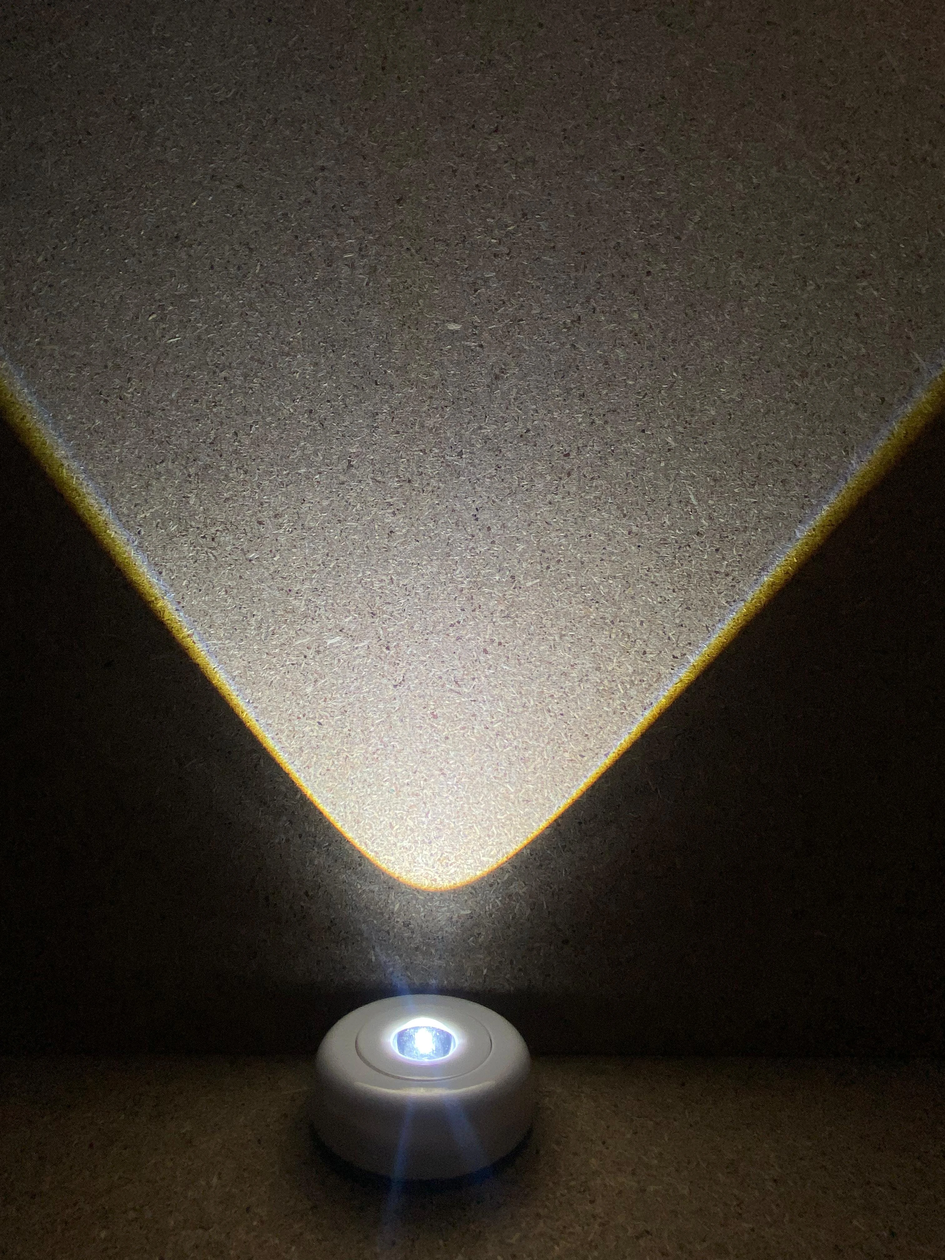 Lámpara LED portátil blanca con luz cálida proyectada en pared, mostrando su haz triangular y base redonda.
