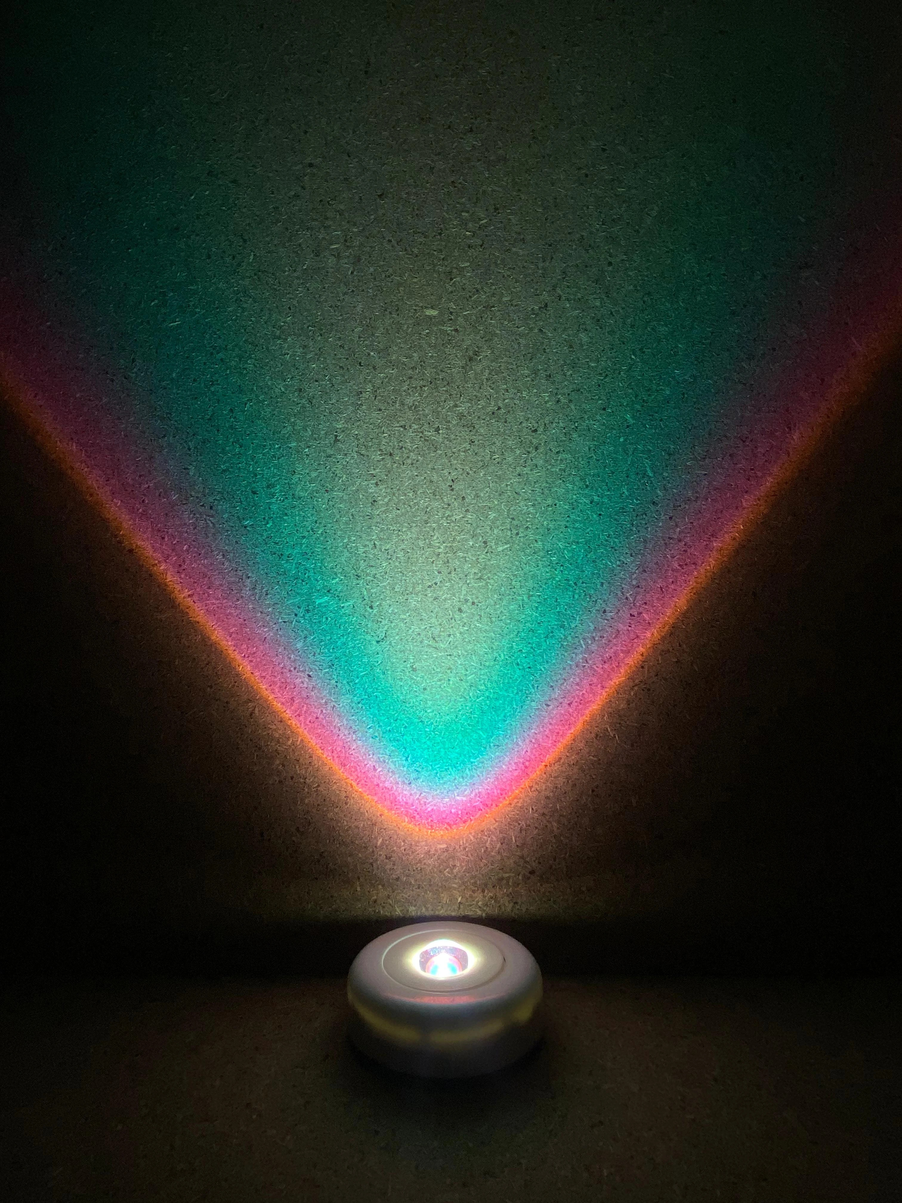 Lámpara LED portátil con batería proyectando luz multicolor en forma de V sobre pared oscura.