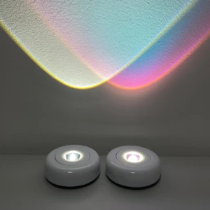 Dos lámparas LED portátiles blancas sobre superficie plana, proyectando luz multicolor en pared texturizada.