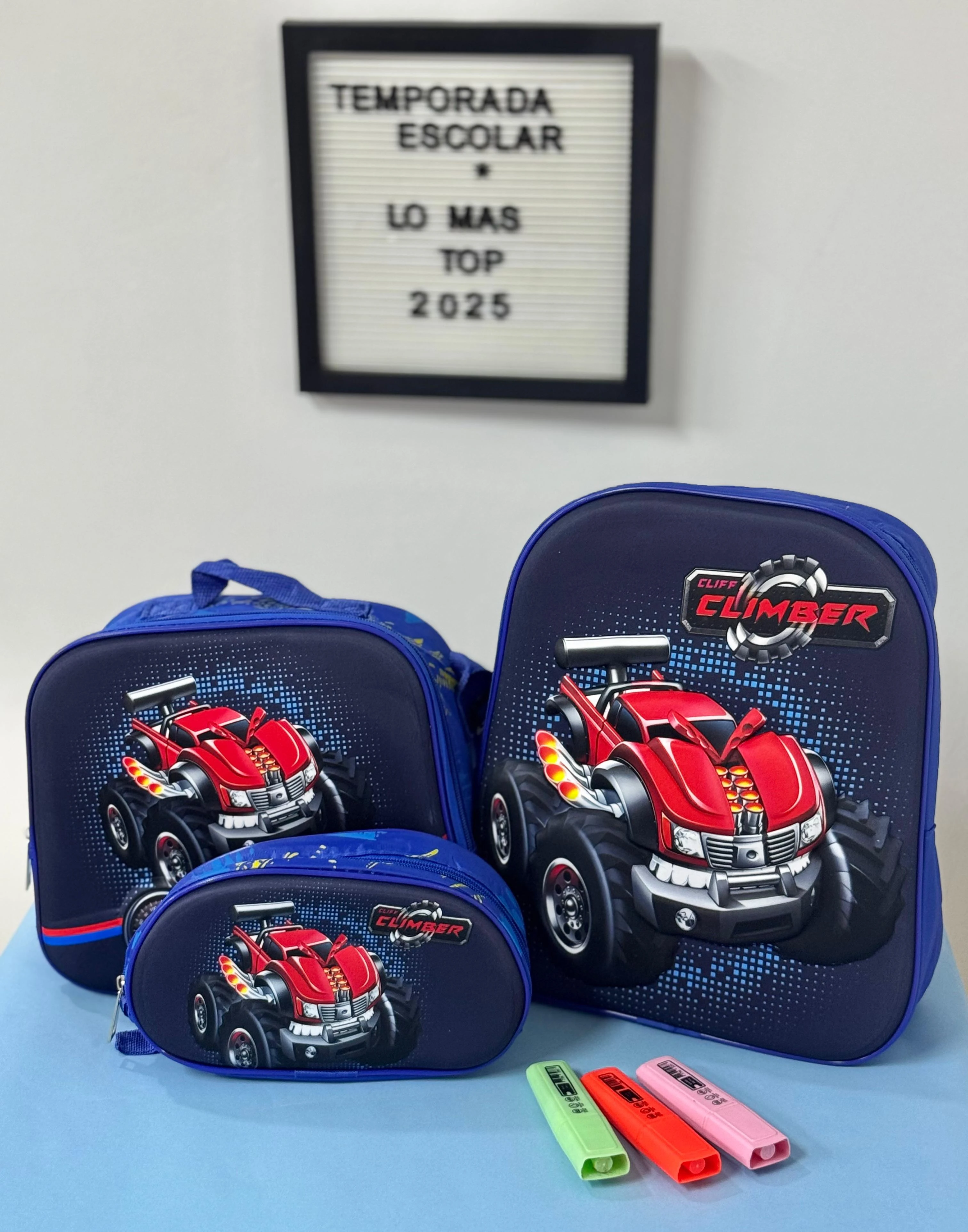 Set de tres piezas de lona azul con diseño 3D de camión rojo, incluye mochila, estuche y almohadilla, acompañado de tres marcadores de colores.