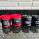 Cuatro vendas de boxeo enrolladas en rojo y negro, con la marca FED y el texto "FIERDOUN SPORT" visible, sobre una superficie oscura brillante.