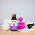 Serux, sérum de colágeno y elastina en frasco ámbar con gotero, caja rosa y toallas blancas sobre superficie de bambú.