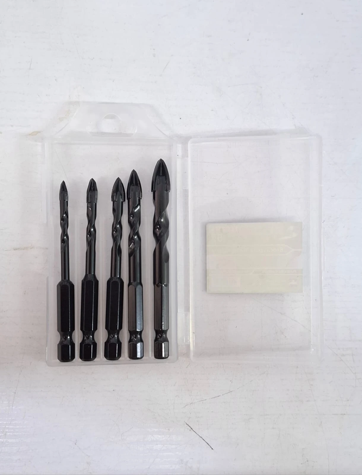 Pack de cuatro brocas multipropósito negras para vidrio y cerámica, con punta de corte y base hexagonal, presentadas en estuche transparente junto a una almohadilla adhesiva.