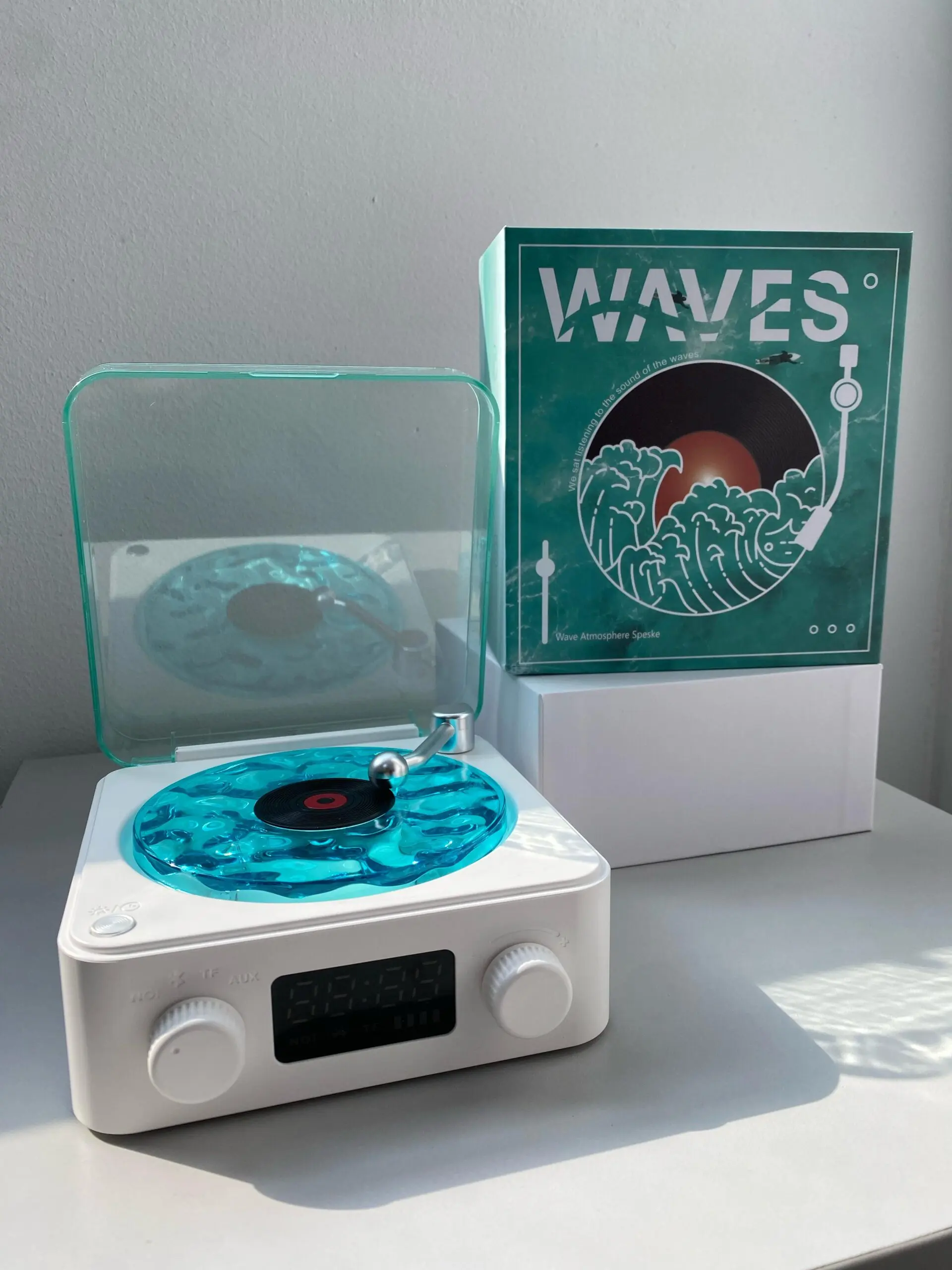 Tocadiscos retro blanco con disco azul translúcido y caja verde con diseño de olas, sobre superficie blanca.