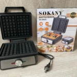 Waflera Sokany SK-08025 de color negro, con tapa abierta mostrando la rejilla interior y panel frontal con perilla de control, junto a su caja de cartón con ilustraciones de uso.