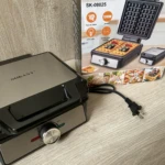 Waflera Sokany SK-08025 de 1200W, color negro y gris metálico, con placa superior abierta y cable enchufado, junto a su caja de cartón mostrando waffles dorados y accesorios incluidos.