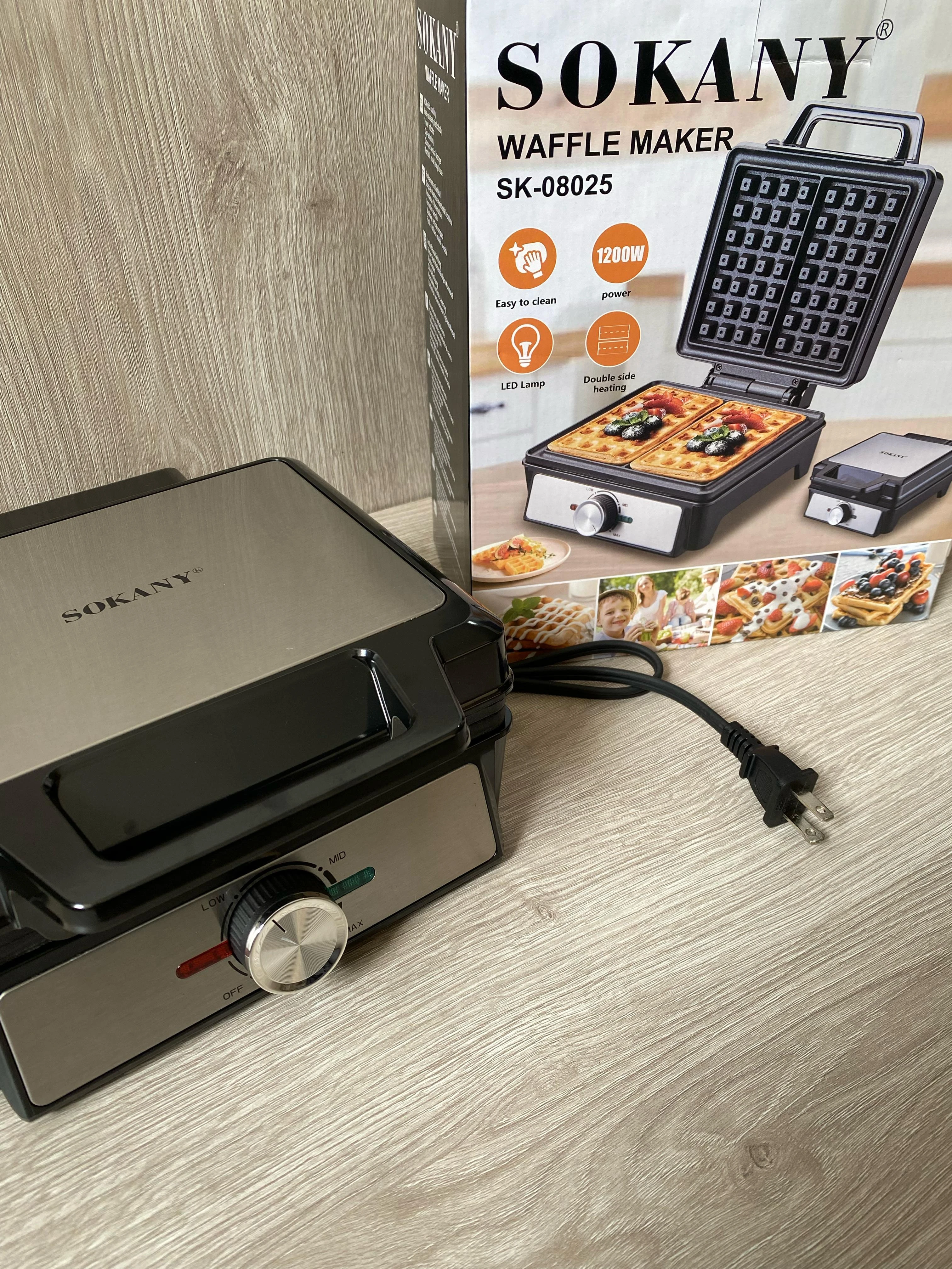 Waflera Sokany SK-08025 de 1200W, color negro y gris metálico, con placa superior abierta y cable enchufado, junto a su caja de cartón mostrando waffles dorados y accesorios incluidos.