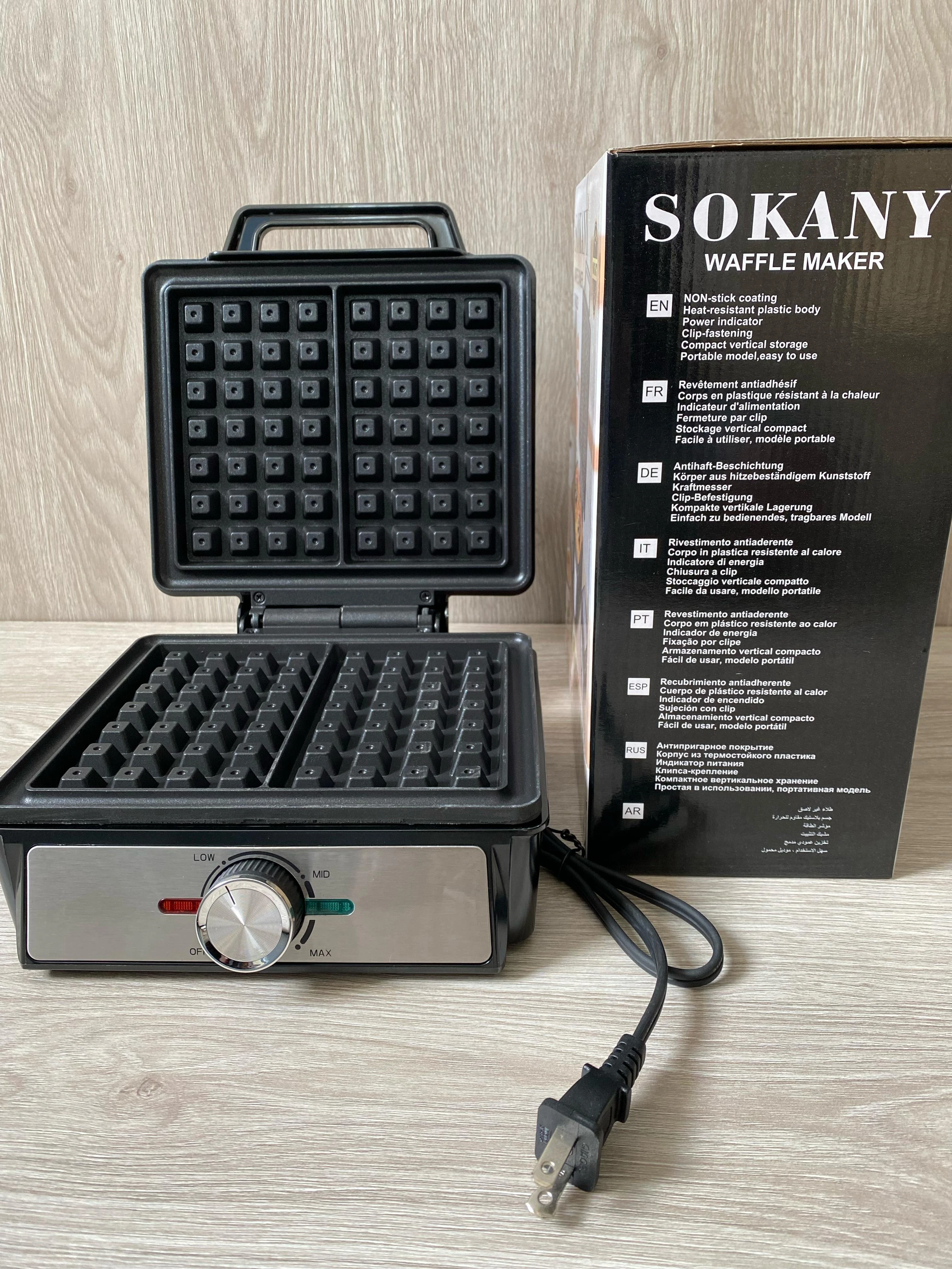 Waflera Sokany de 1200W, color negro, con tapa abierta mostrando placas de cocción y panel frontal con perilla de control, junto a su caja original.