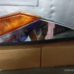 Indicador lateral compacto con carcasa cromada y lente ámbar, colocado sobre cajas de cartón.