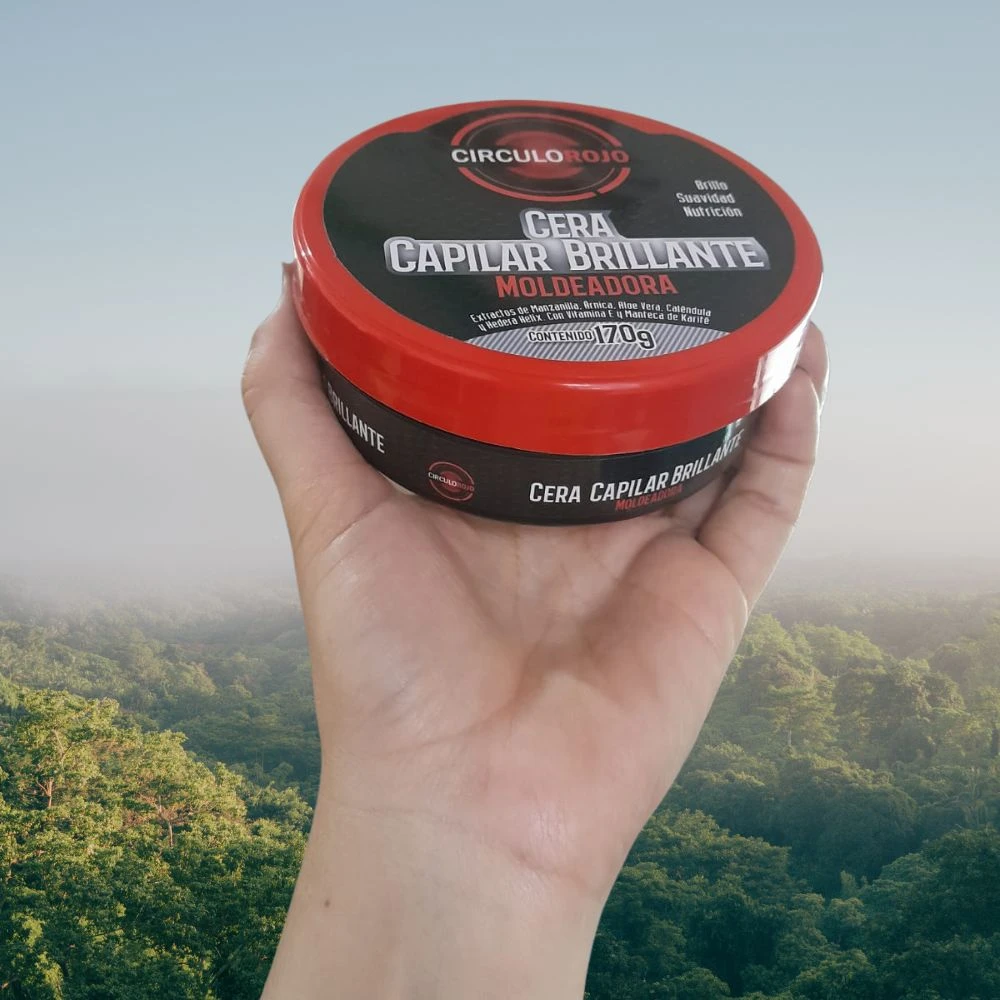 Contenedor redondo de cera capilar moldeadora con karité y aloe vera, color rojo brillante, etiqueta negra con texto blanco y detalles en dorado, mostrando peso neto de 170g, sostenido en mano sobre fondo de bosque neblinoso.