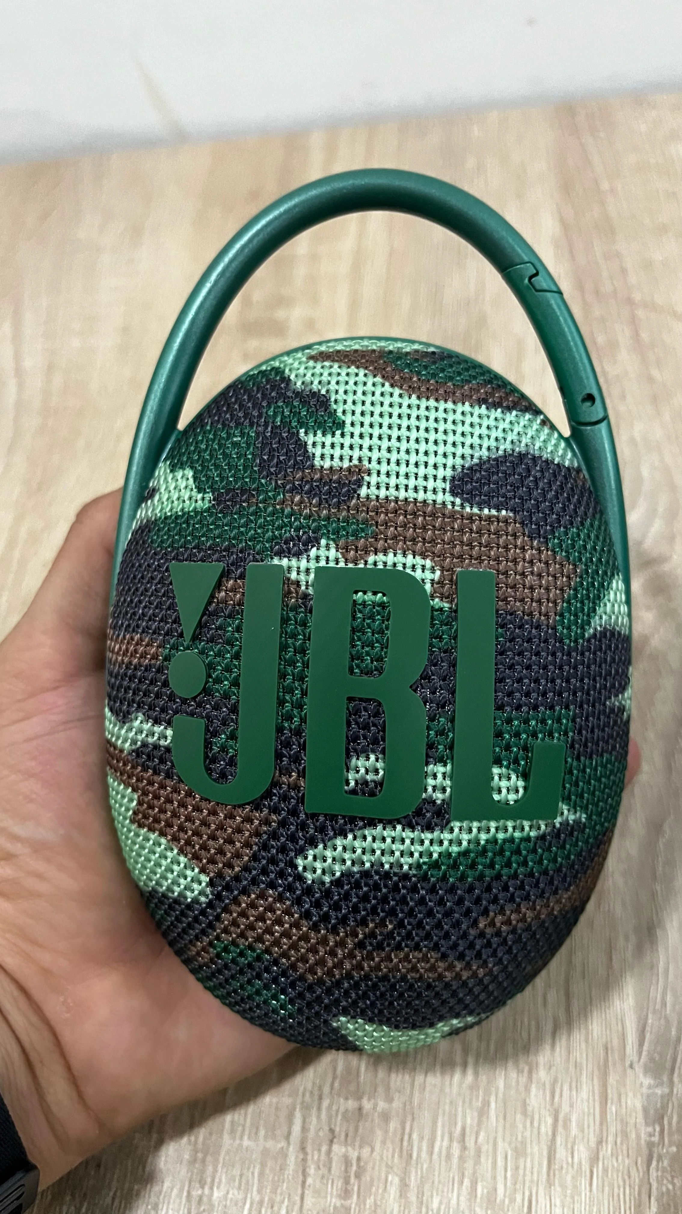 Parlante JBL Clip 5 en patrón camuflaje verde, sostenido en mano, con asa integrada y logotipo visible.