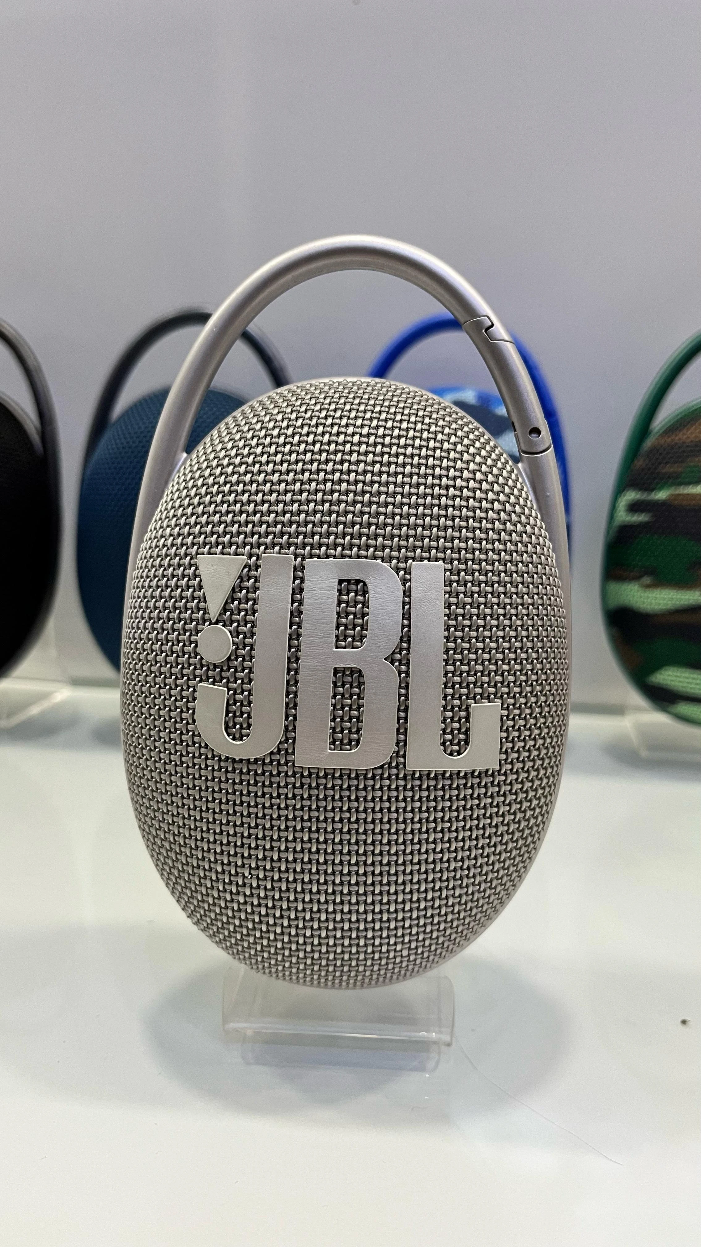 Parlante JBL Clip 5 en color gris, con textura de malla y logo frontal visible, sobre soporte transparente.