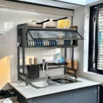 Platillera escurridora de aluminio con tapa protectora, color gris oscuro, instalada sobre fregadero de cocina con vajilla y utensilios organizados.