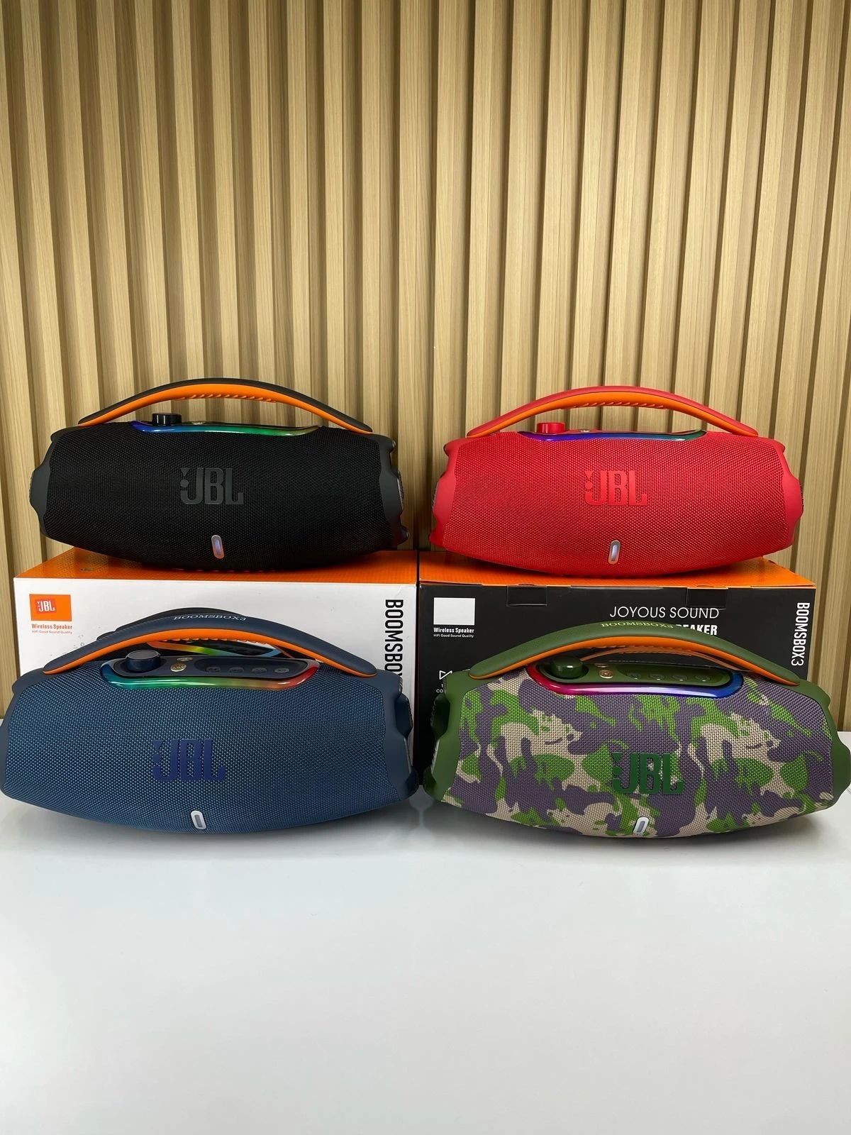 Cuatro altavoces portátiles JBL Boombox 3 en colores negro, rojo, azul y camuflaje, sobre sus cajas originales, con asas ergonómicas y acabado mate.