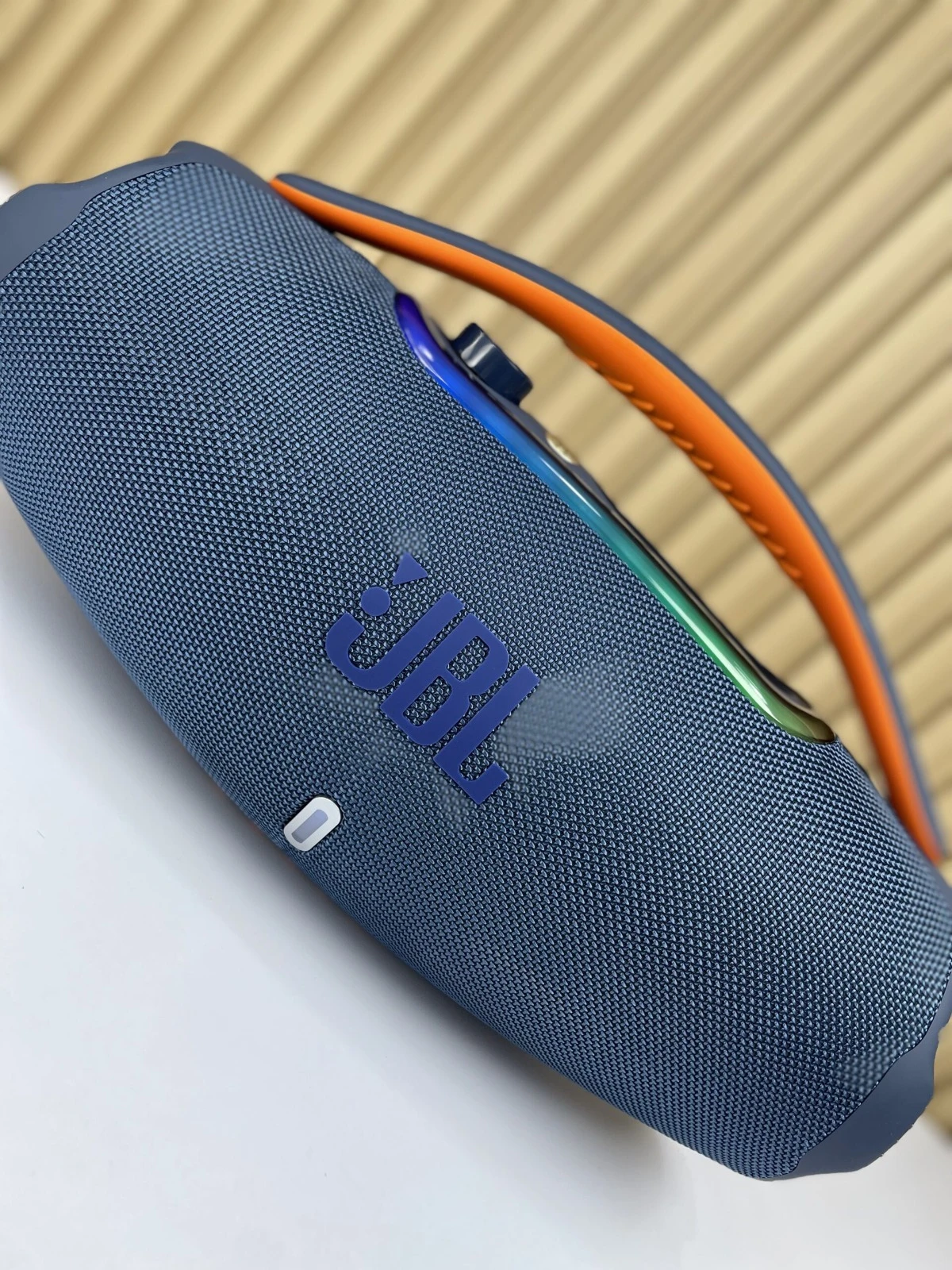 Altavoz portátil JBL Boombox 3 en color azul marino con textura de malla, asa naranja y detalles multicolores en la parte superior.