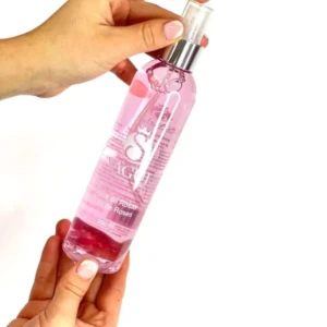 Botella de agua de rosas Sagu con ácido láctico y aceite de rosa Irazu, presentada en un frasco transparente con líquido rosado y etiqueta blanca, sostenida por dos manos sobre fondo blanco.