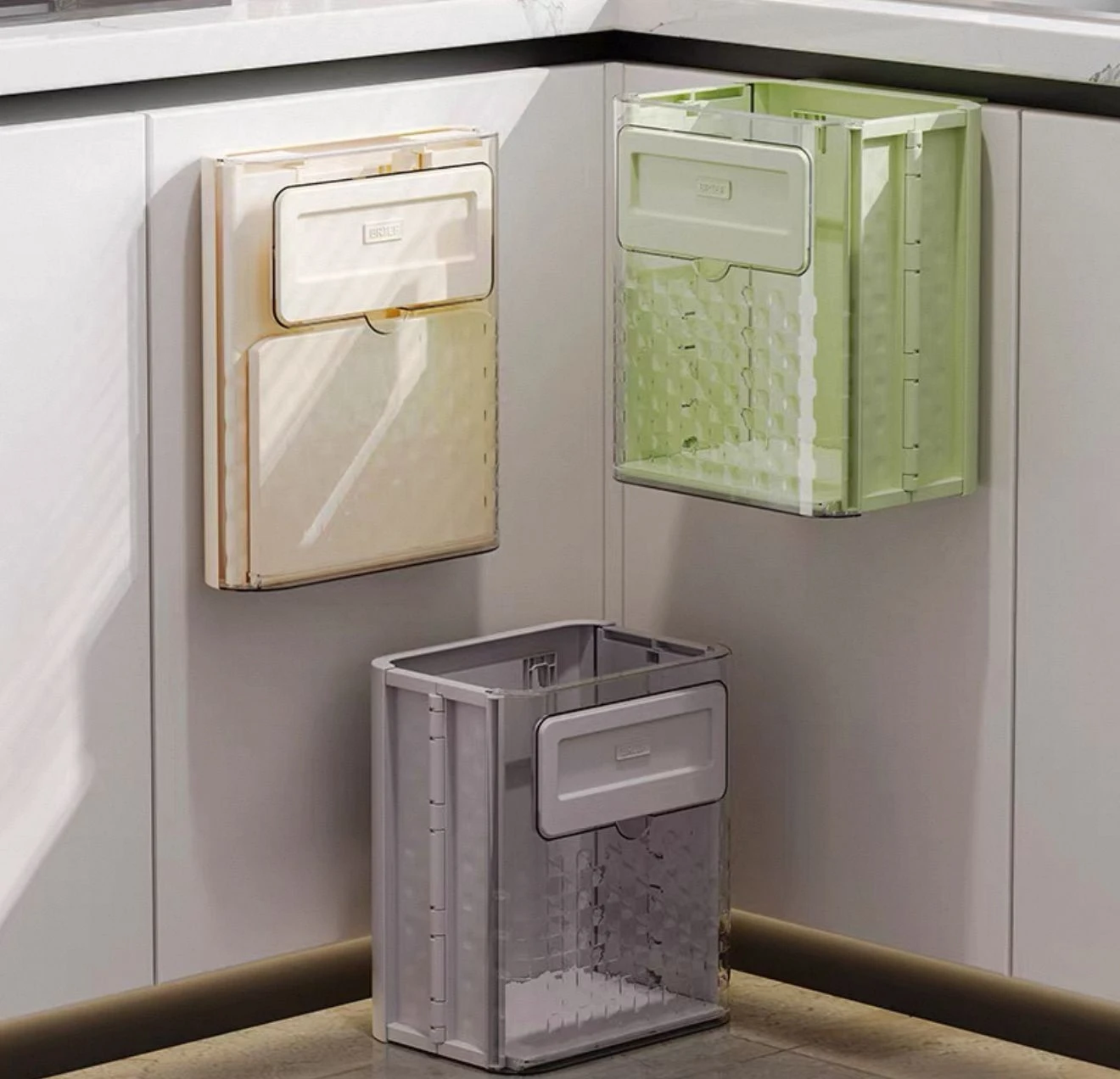 Tres basureros plegables de plástico transparente en colores beige, verde y gris, montados en pared y uno sobre encimera, con tapa frontal y diseño compacto para espacios reducidos.