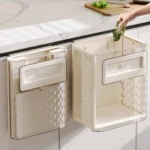 Basurero plegable beige con tapa y compartimento inferior, montado en gabinete de cocina; mano depositando residuos verdes.