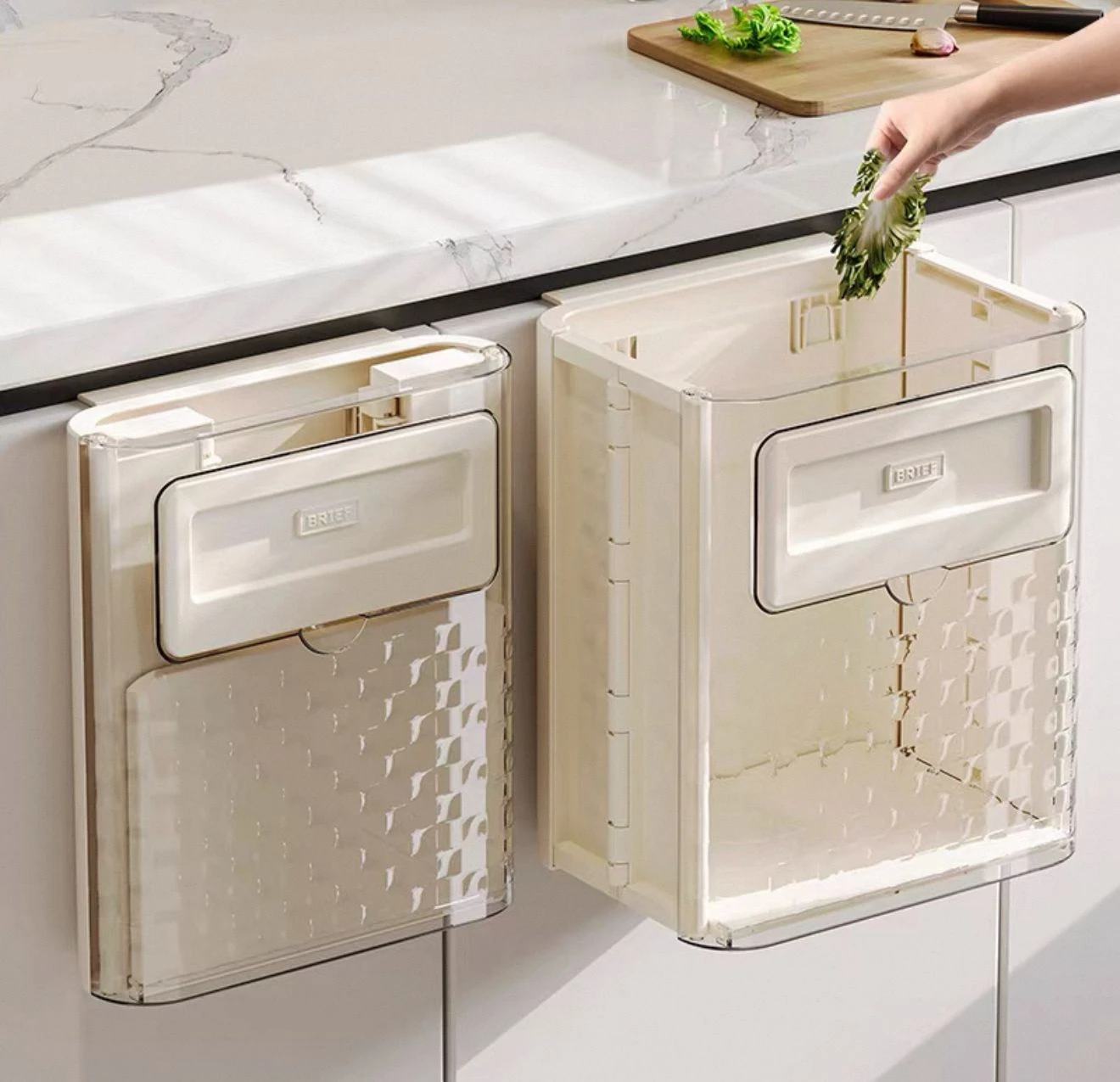 Basurero plegable beige con tapa y compartimento inferior, montado en gabinete de cocina; mano depositando residuos verdes.