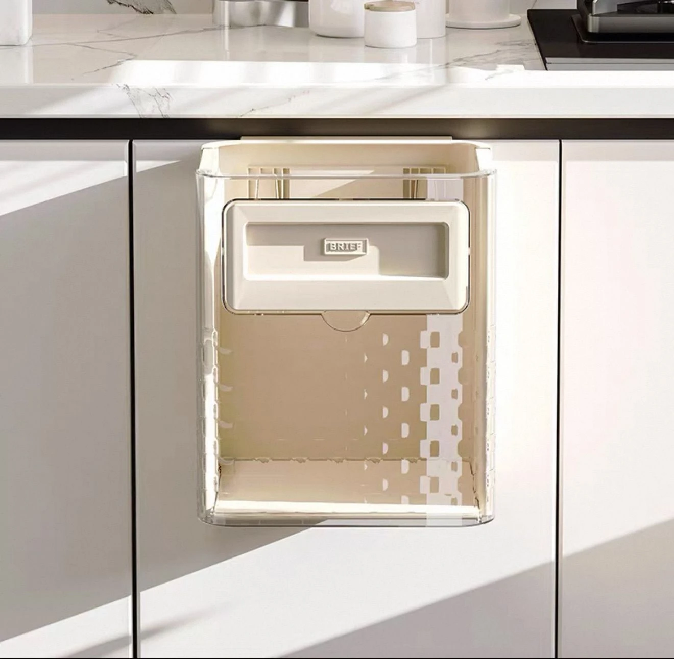 Basurero plegable beige con tapa blanca, montado en puerta de gabinete blanco. Diseño compacto con estructura transparente y panel decorativo. Ideal para cocina.