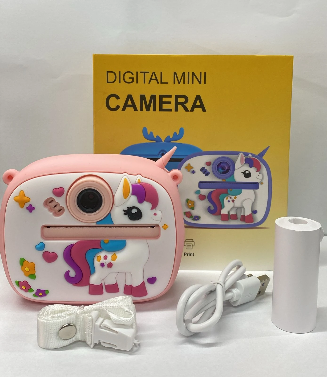 Cámara digital mini infantil rosa con diseño de unicornio, incluye cinta de papel, cable USB y estuche. Caja amarilla con del producto.