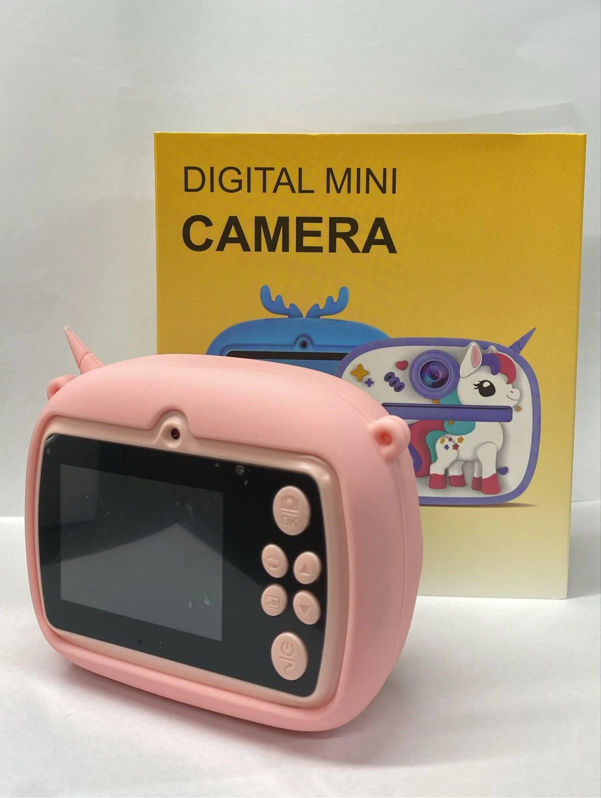 Cámara digital mini rosa con pantalla frontal y botones laterales, sobre caja amarilla que muestra de unicornio.