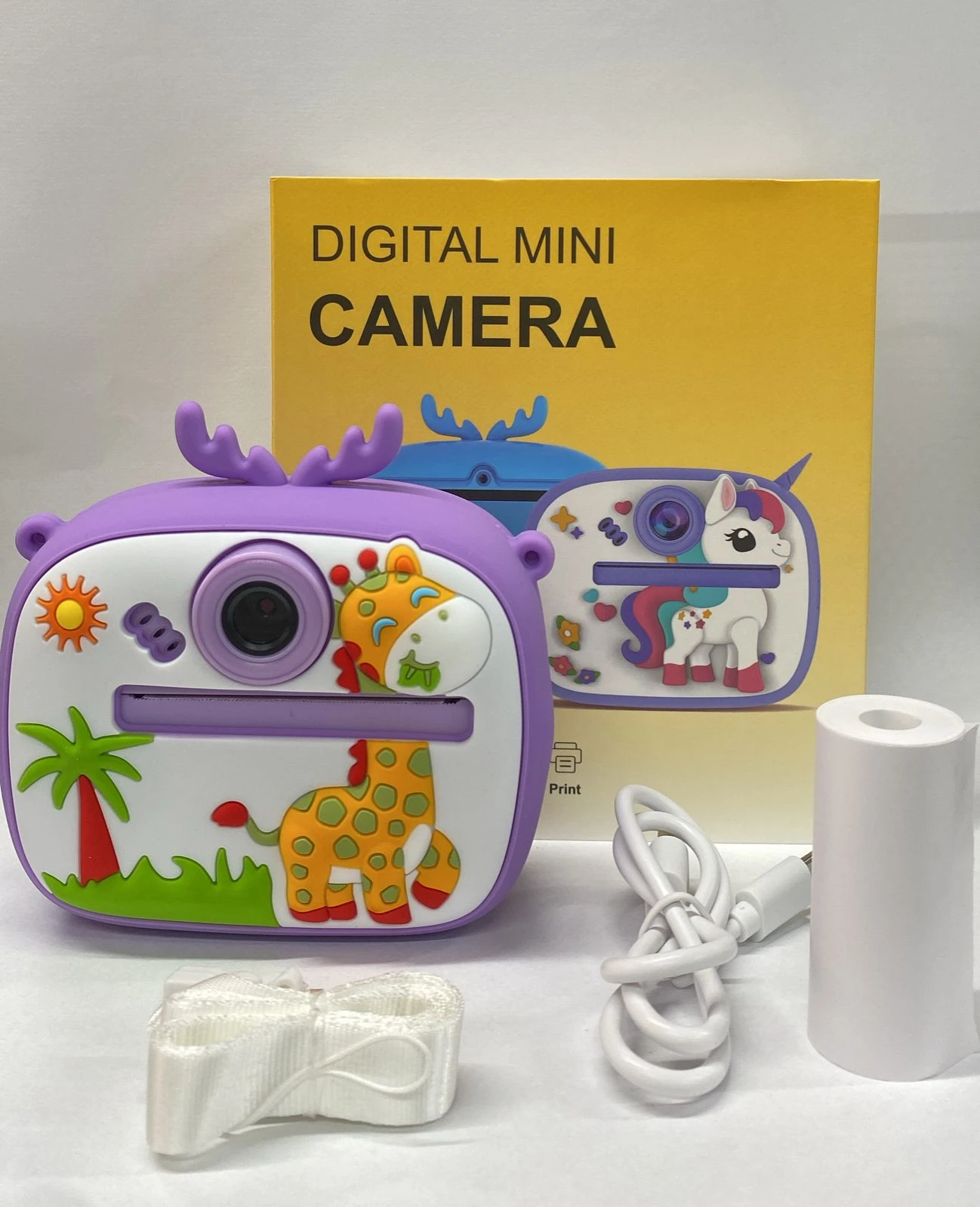 Cámara digital mini infantil en color lila con temática de unicornio y jirafa, incluye cinta de impresión, cable USB y estuche de transporte.